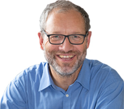 Ein lächelnder weißer Mann in einem blauen Hemd mit grauem Haar und grauer Brille.